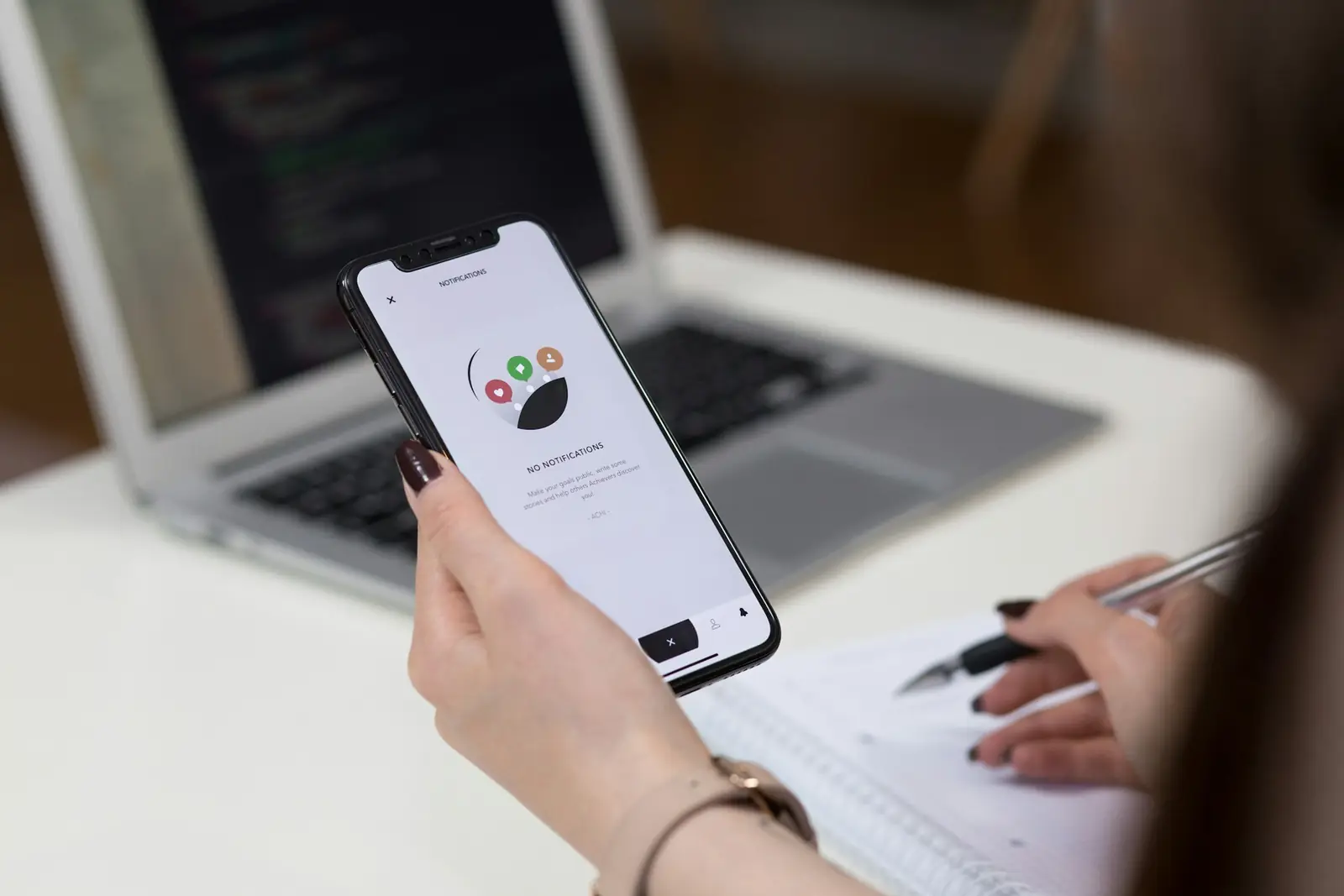 Pessoa segura um celular com um aplicativo aberto ao lado de um notebook e de um caderno de anotações
