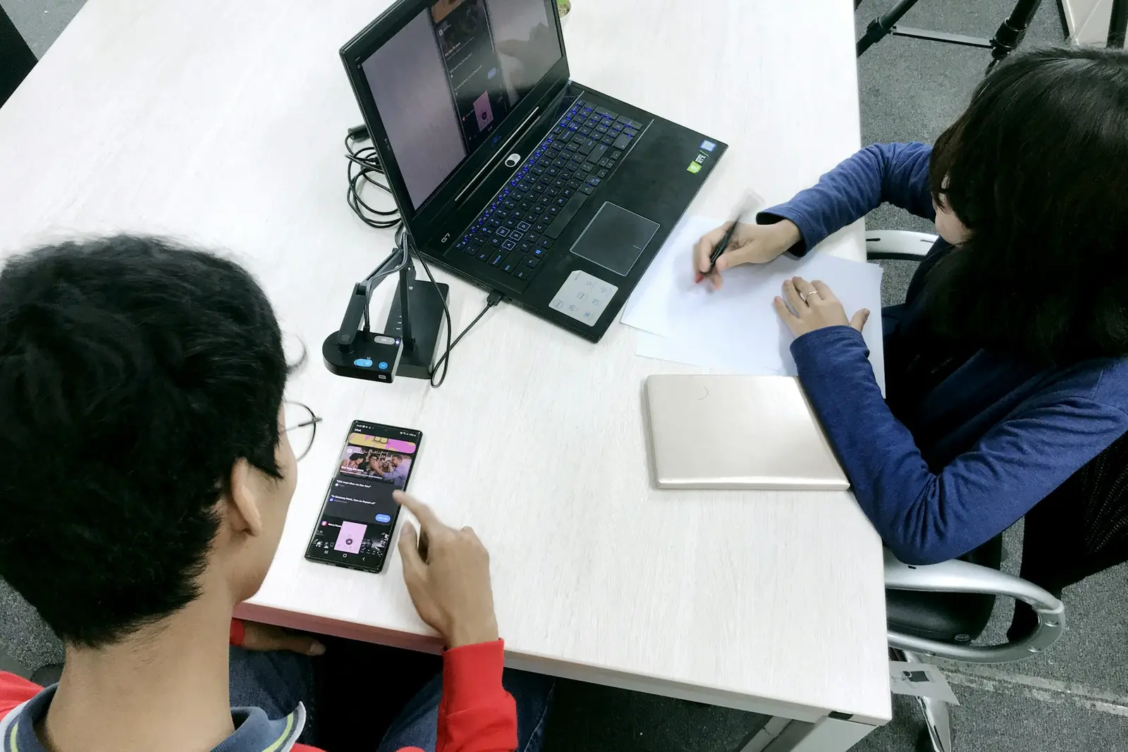 Duas pessoas numa mesa branca: uma testa um aplicativo no celular enquanto a outra observa e anota, com um notebook ao lado